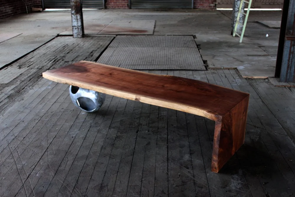 A live-edge wooden bench balanced on a large round polished metal sphere, set on a worn wooden floor inside an industrial studio space with warm brown and gray tones