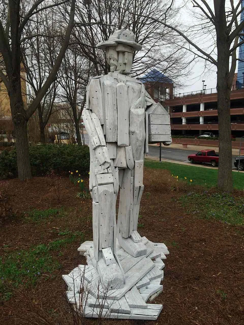 Weathered wooden sculpture of a standing figure in a hat, set among mulch and garden greenery, in muted brown and gray tones